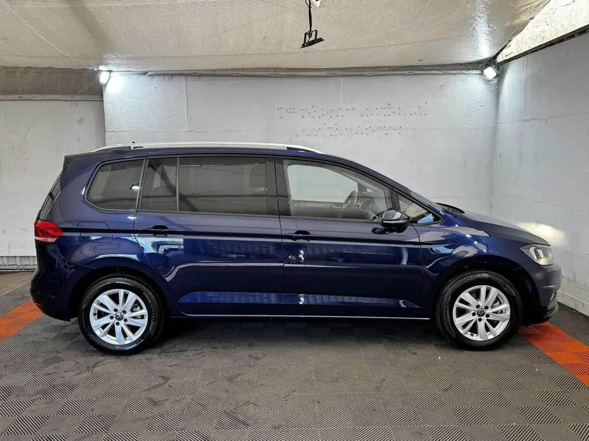 Vue de profil côté gauche d'un Volkswagen Touran bleu 2024 stationné en intérieur sur sol carrelé.