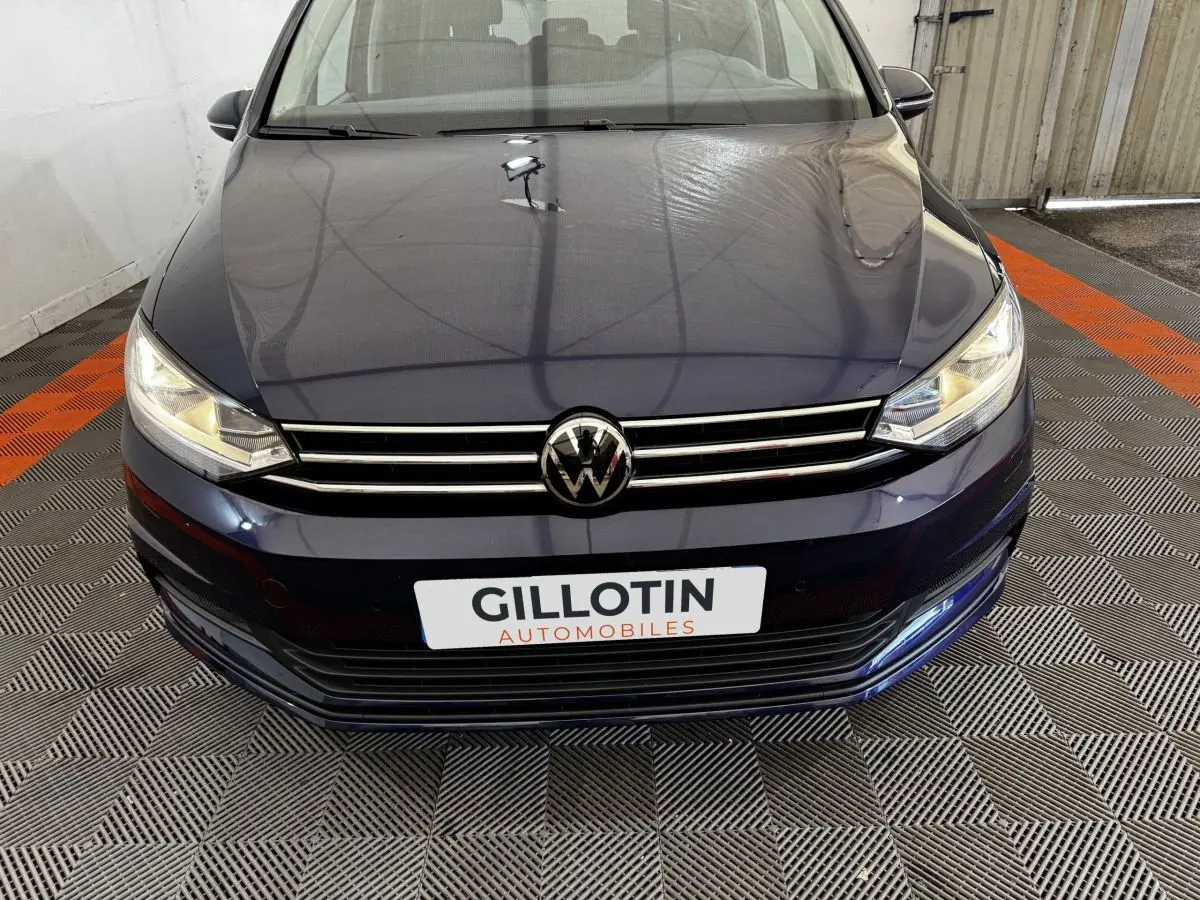 Vue avant rapprochée d’un Volkswagen Touran bleu 2024 avec calandre chromée et phares allumés dans un garage.