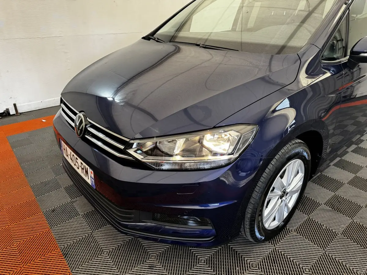 Vue 3/4 avant droit d'un Volkswagen Touran bleu 2024 avec phares allumés sur sol à damier gris et orange.