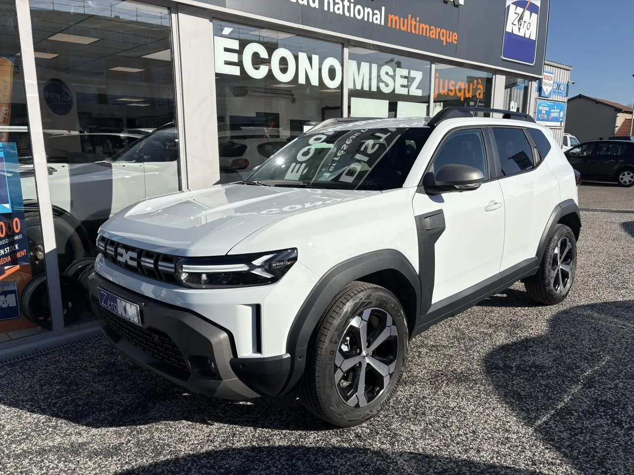 SUV Dacia Duster blanc métallisé en 3/4 avant droit, avec barres de toit noires et jantes alliage 18 pouces.