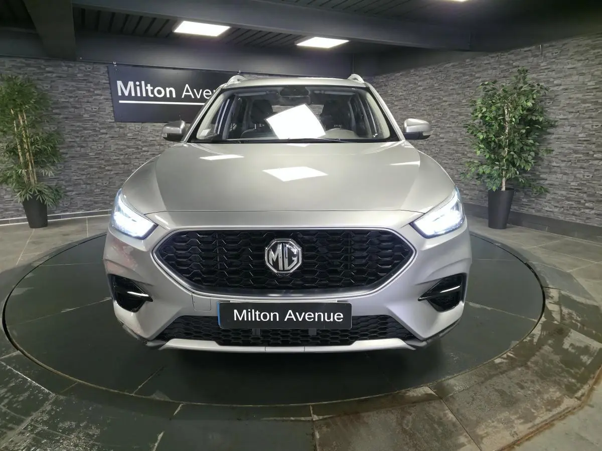 Vue avant du MG ZS gris métallisé 2023 avec calandre noire hexagonale et phares LED allumés.