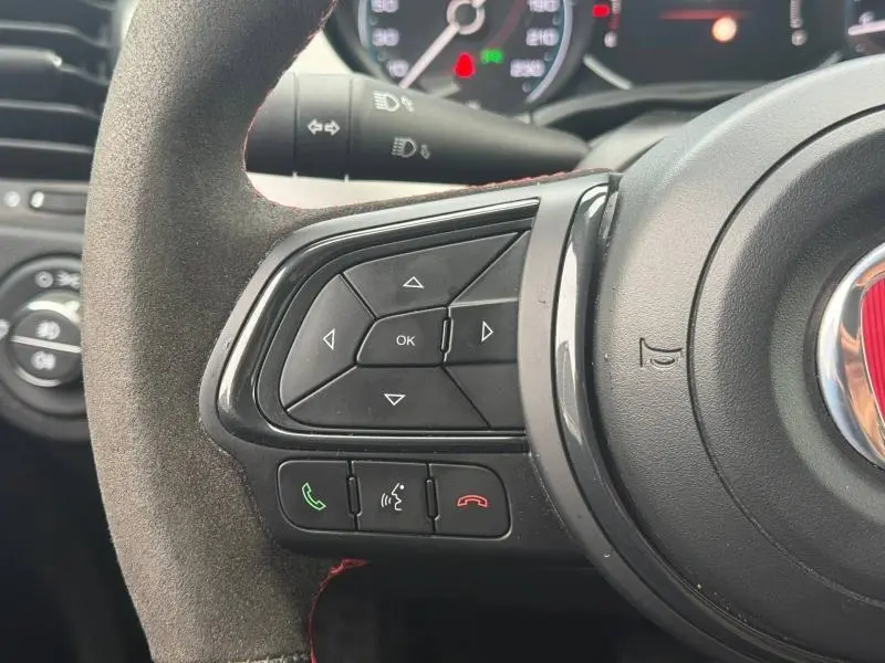 Gros plan sur les commandes gauche du volant en alcantara noir avec surpiqûres rouges du Fiat 500X Rouge Passione pastel 2024.