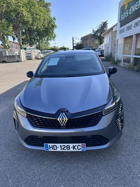 Vue avant d'une Renault Clio gris métallisé 2025, avec calandre noire et phares LED allumés, stationnée en extérieur.