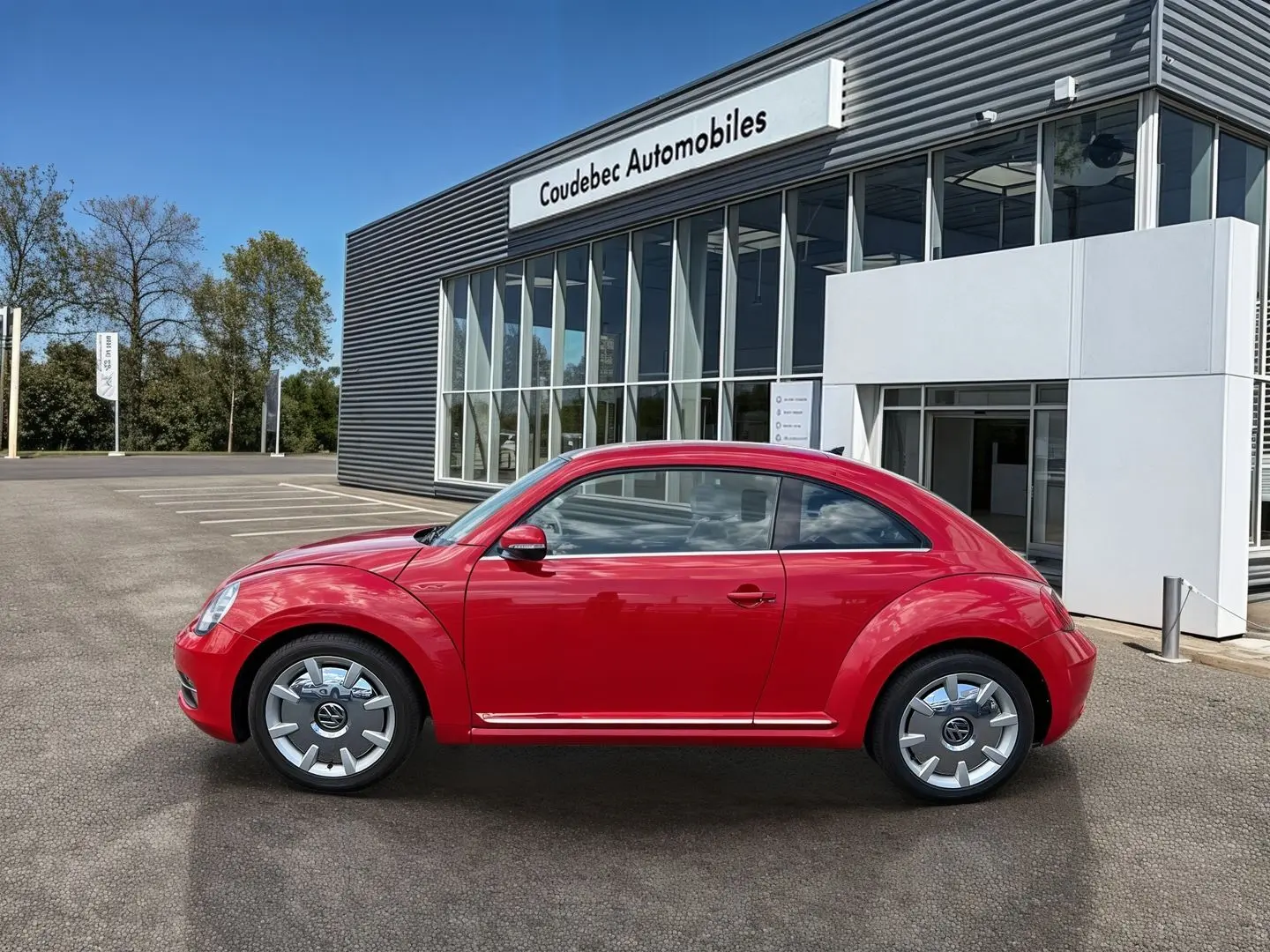 Profil droit d'une Volkswagen Coccinelle rouge Tornade 2012, avec jantes 18 pouces et carrosserie brillante devant un concessionnaire.