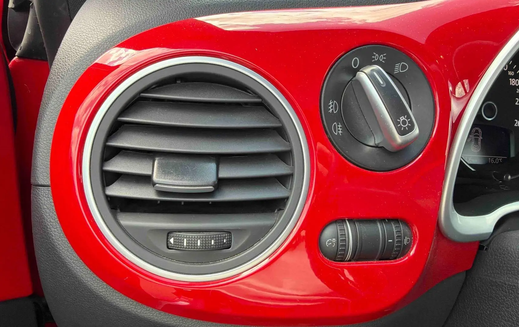 Détail du tableau de bord rouge brillant côté gauche avec commande des phares et bouche d’aération ronde sur Volkswagen Coccinelle 2012.