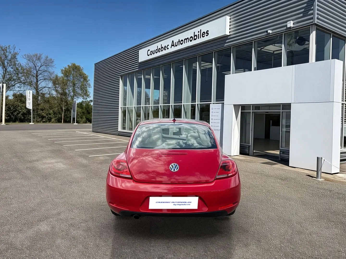 Vue arrière d'une Volkswagen Coccinelle rouge Tornade garée devant un bâtiment moderne de concession automobile.