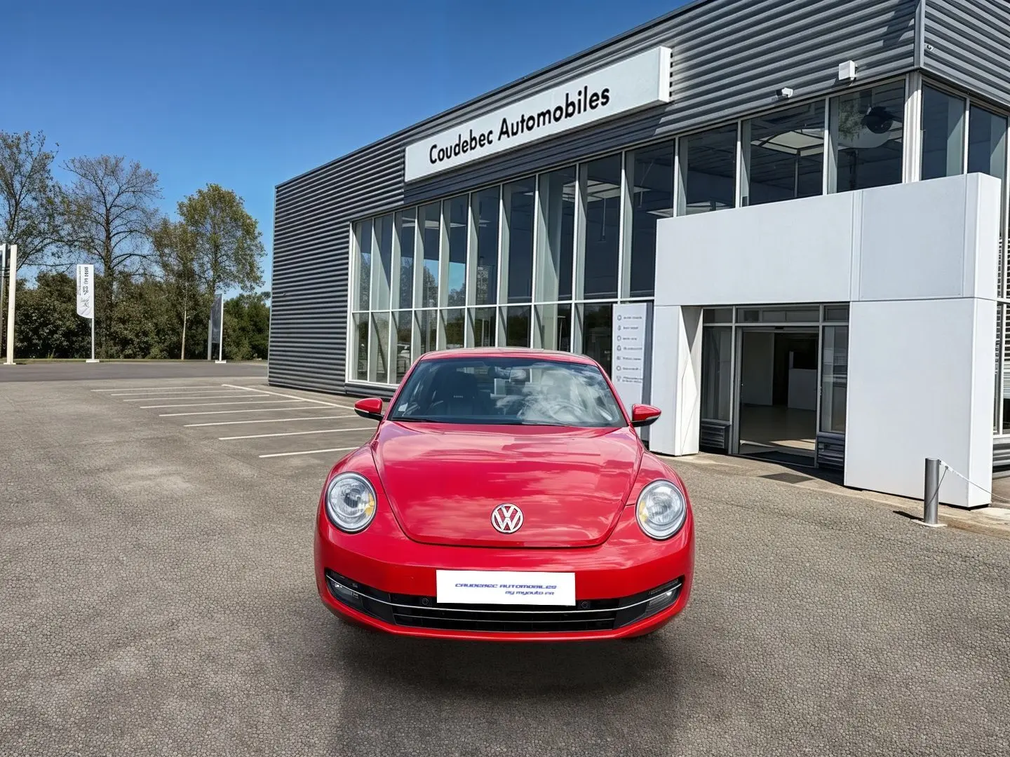 Vue de face d'une Volkswagen Coccinelle rouge Tornade garée devant un bâtiment moderne de concession automobile.