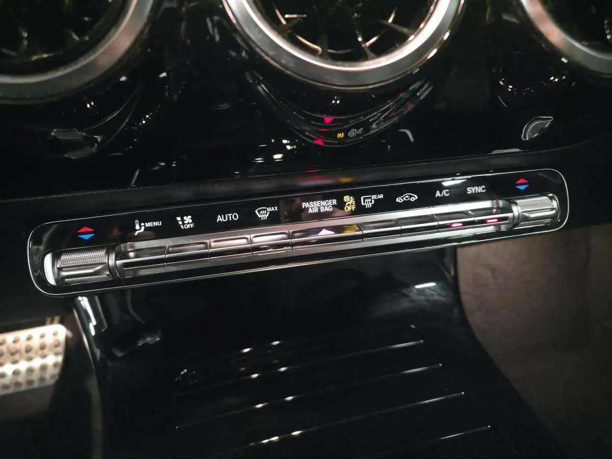 Gros plan sur la console centrale noire brillante avec commandes de climatisation digitale et buses d’aération rondes chromées.