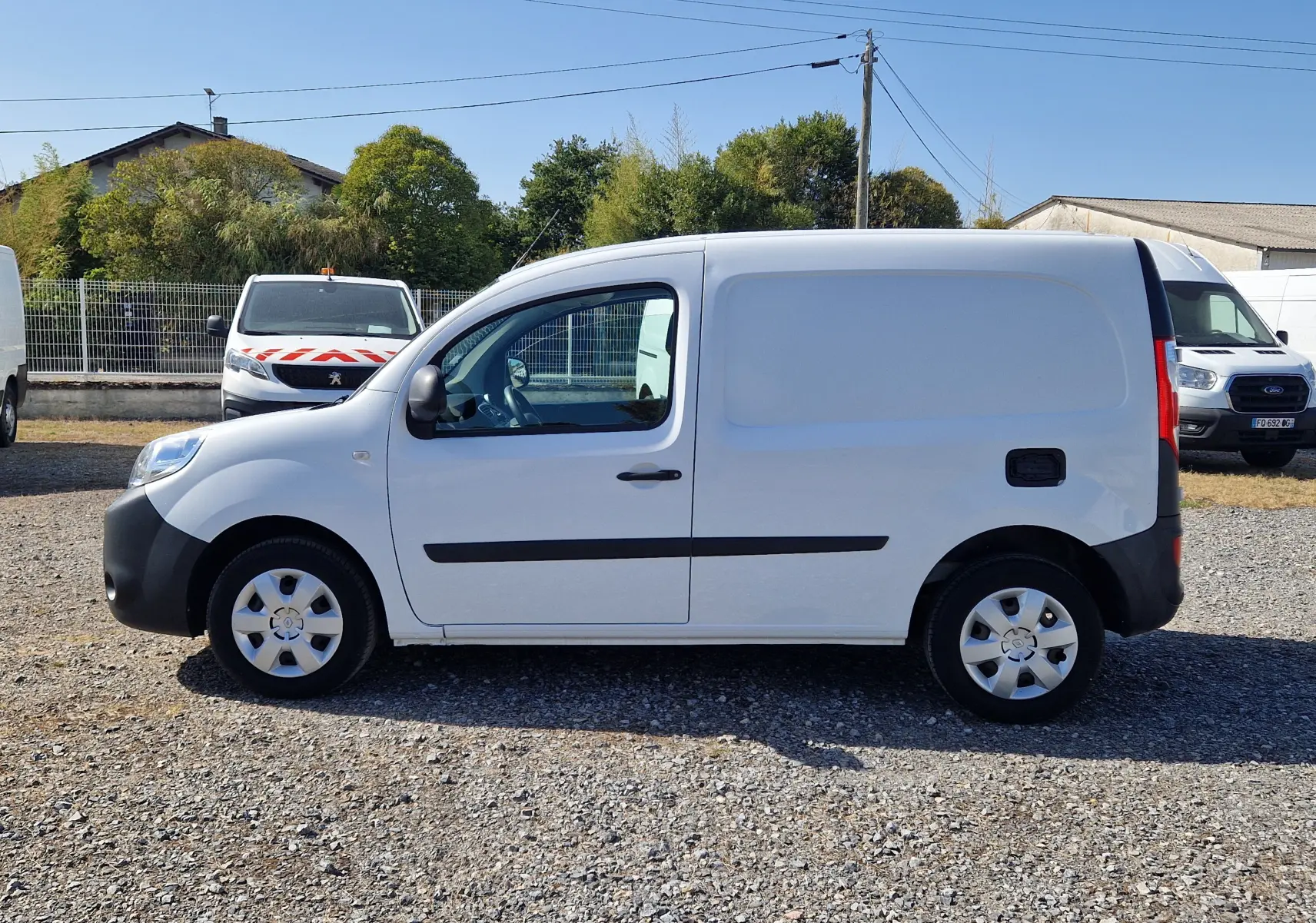 Vue de profil côté gauche d'un Renault Kangoo blanc utilitaire avec porte latérale pleine, stationné sur un sol gravillonné.