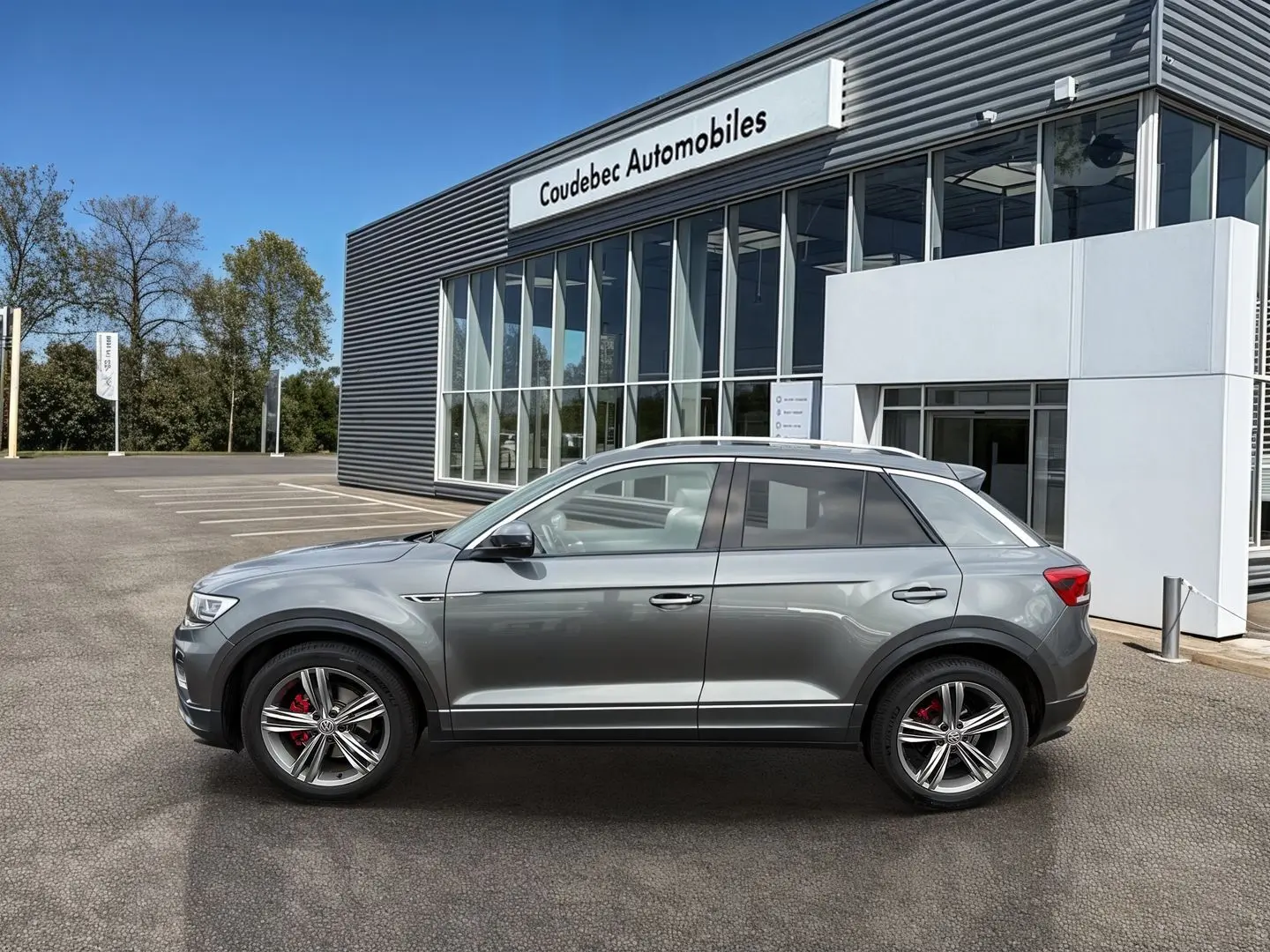 Profil côté gauche d'un Volkswagen T-Roc gris Indium Metal avec jantes alliage et étriers de frein rouges, devant un bâtiment.