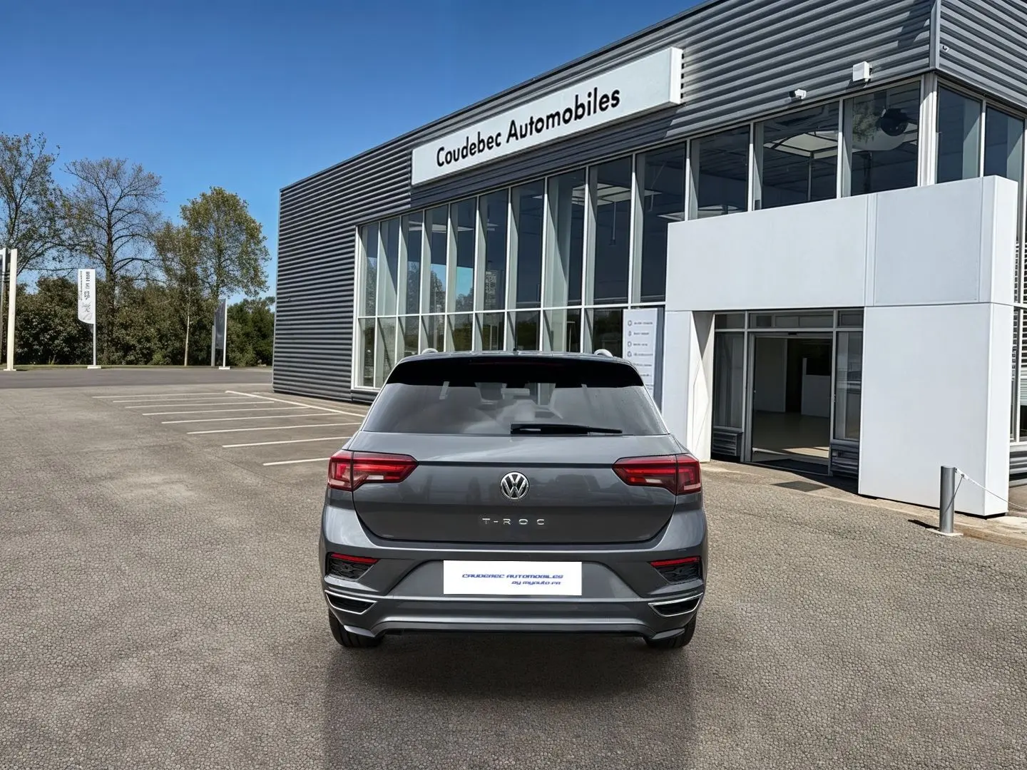 Vue arrière d'un Volkswagen T-Roc gris Indium Metal garé devant un bâtiment moderne de concession automobile.