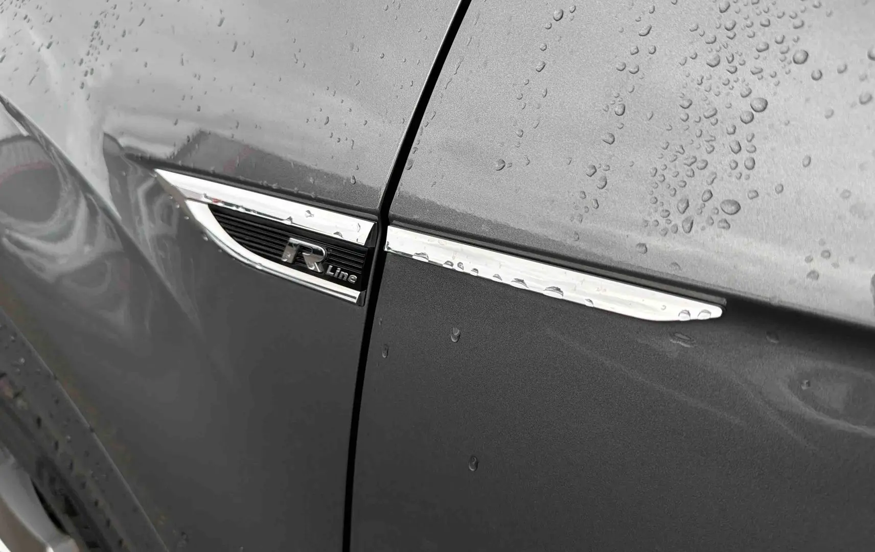 Gros plan sur l’aile avant gauche gris Indium métallisé du Volkswagen T-Roc avec logo R-Line et gouttes de pluie.