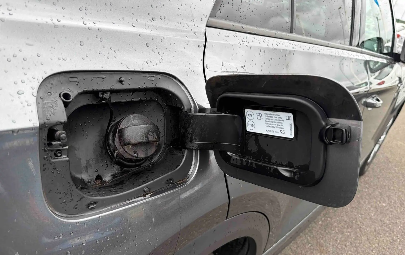 Vue rapprochée du volet de remplissage essence ouvert côté gauche d’un Volkswagen T-Roc gris Indium Metal sous la pluie.