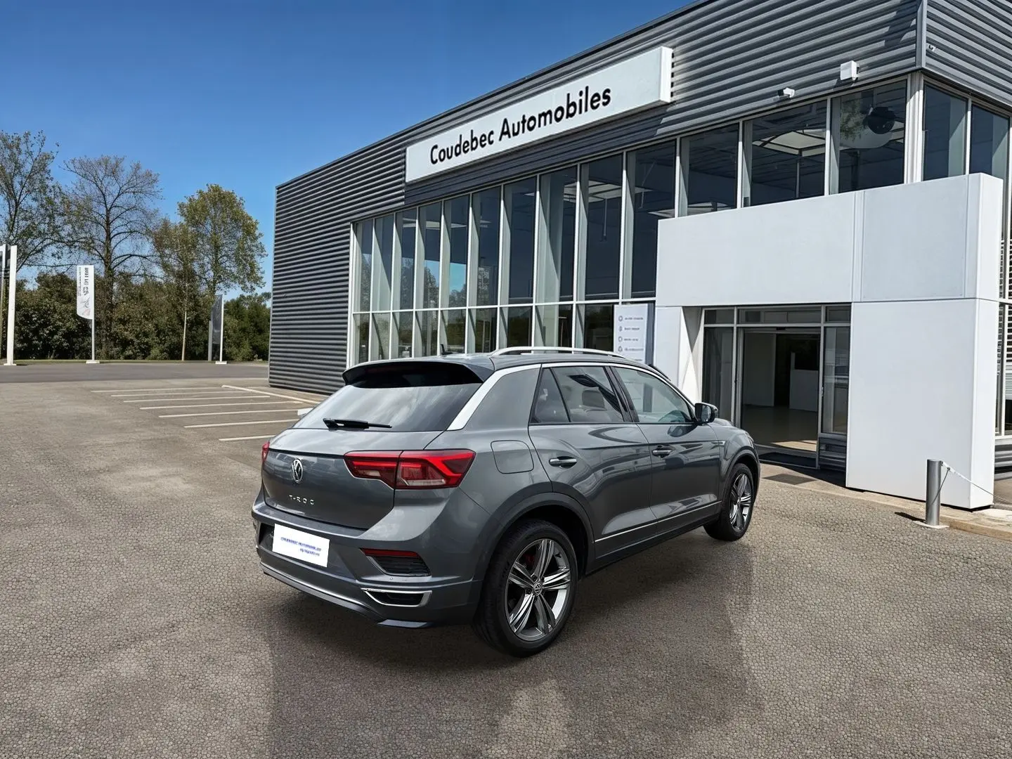 Volkswagen T-Roc gris Indium métal vu en 3/4 arrière droit devant un concessionnaire Coudebec Automobiles.
