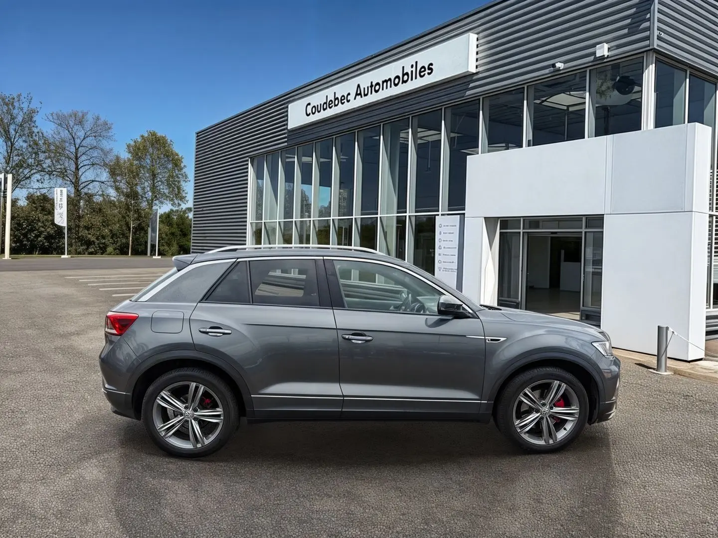 Vue latérale droite d'un Volkswagen T-Roc gris Indium Metal avec jantes alliage et étriers rouges, devant un bâtiment commercial.