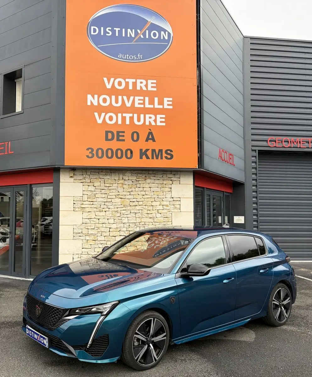 Peugeot 308 bleu 3/4 avant droit, avec feux LED et jantes alliage, devant un bâtiment commercial.