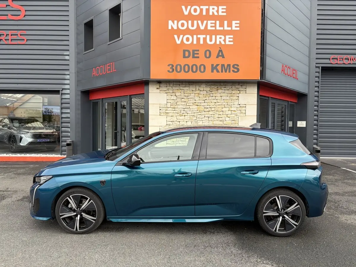 Profil latéral gauche d'une Peugeot 308 bleu métallisé 2025 garée devant un bâtiment avec enseigne orange.