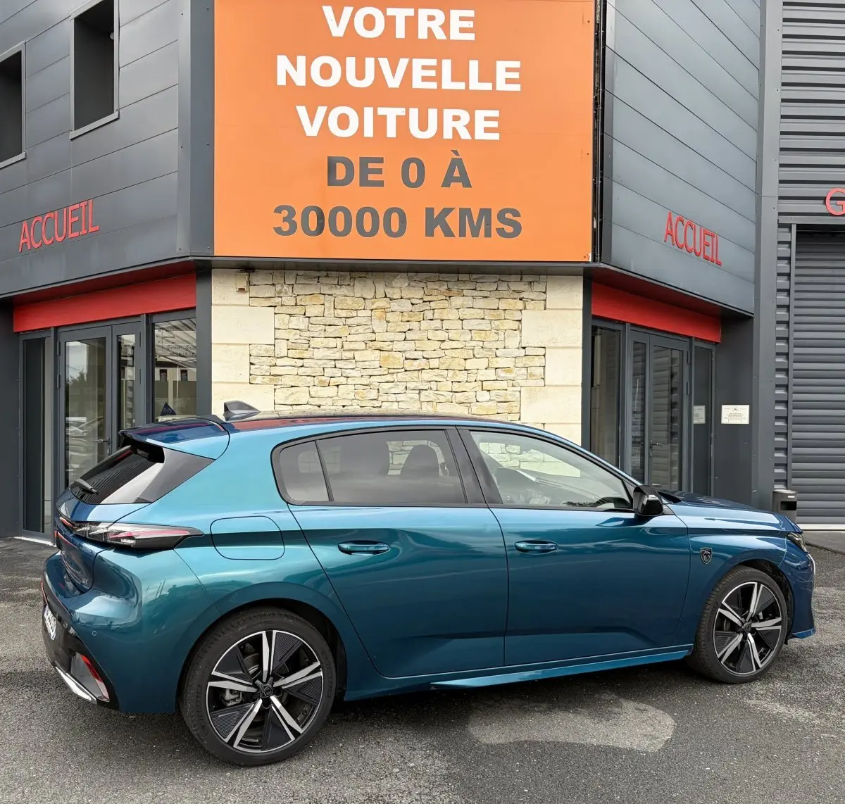 Vue latérale droite d'une Peugeot 308 1.2 Hybrid 145 GT bleu métallisé, garée devant un bâtiment avec enseigne orange.