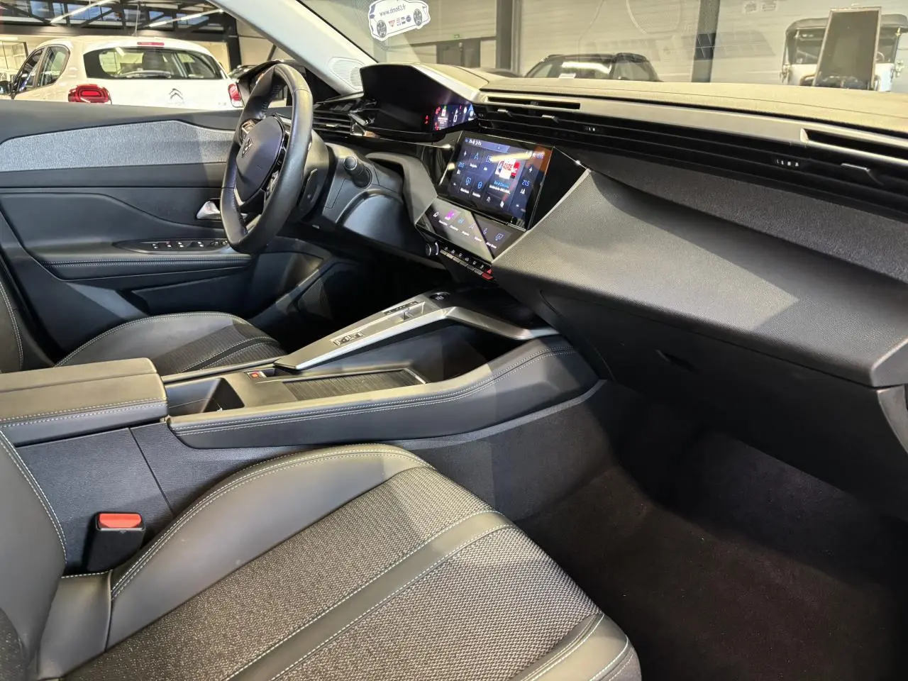 Vue intérieure côté conducteur de la Peugeot 308 SW 2024, sellerie tri matières noire avec surpiqûres bleues et tableau de bord moderne.