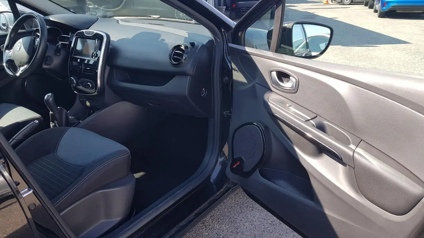Intérieur avant gauche d’une Renault Clio IV noire, montrant tableau de bord, volant cuir et porte ouverte.