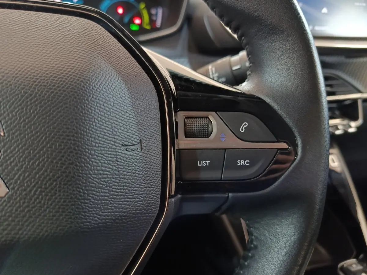 Gros plan sur les commandes droite du volant cuir noir du Peugeot 208 électrique blanc, avec tableau de bord en arrière-plan flou.