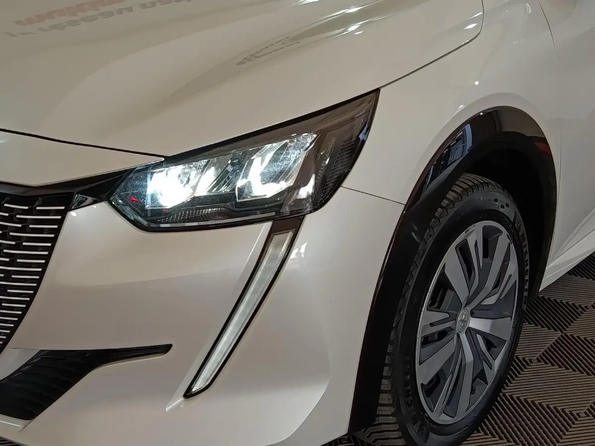 Gros plan sur l'avant droit blanc d'une Peugeot 208 électrique avec phares LED allumés et jante stylisée.