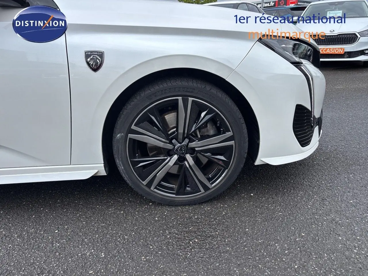 Gros plan sur la roue avant droite et l'aile avant d'une Peugeot 308 SW blanche nacrée, avec logo lion visible.