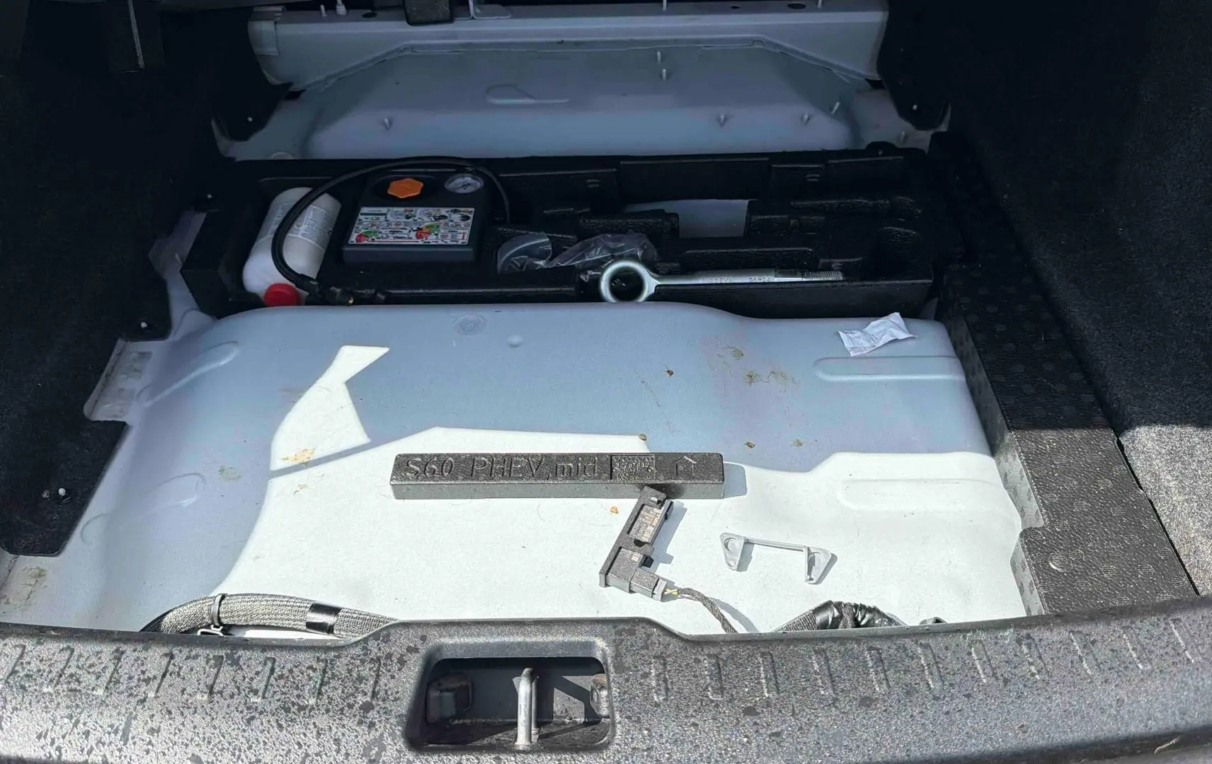 Vue du coffre ouvert d'une Volvo S60 blanche montrant le compartiment de rangement sous plancher avec outils et kit de réparation.