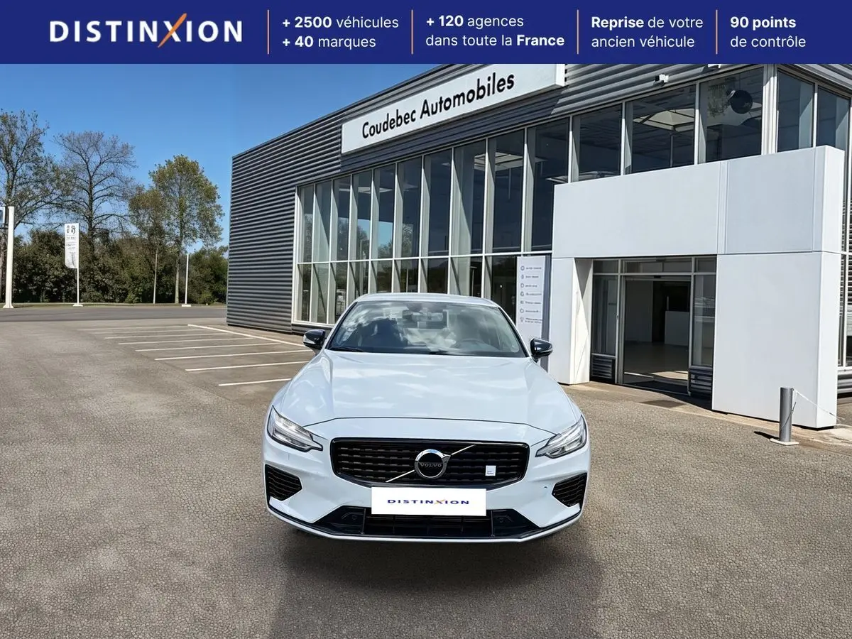 Vue avant d'une Volvo S60 blanche avec calandre noire brillante et feux LED, stationnée devant un bâtiment commercial.