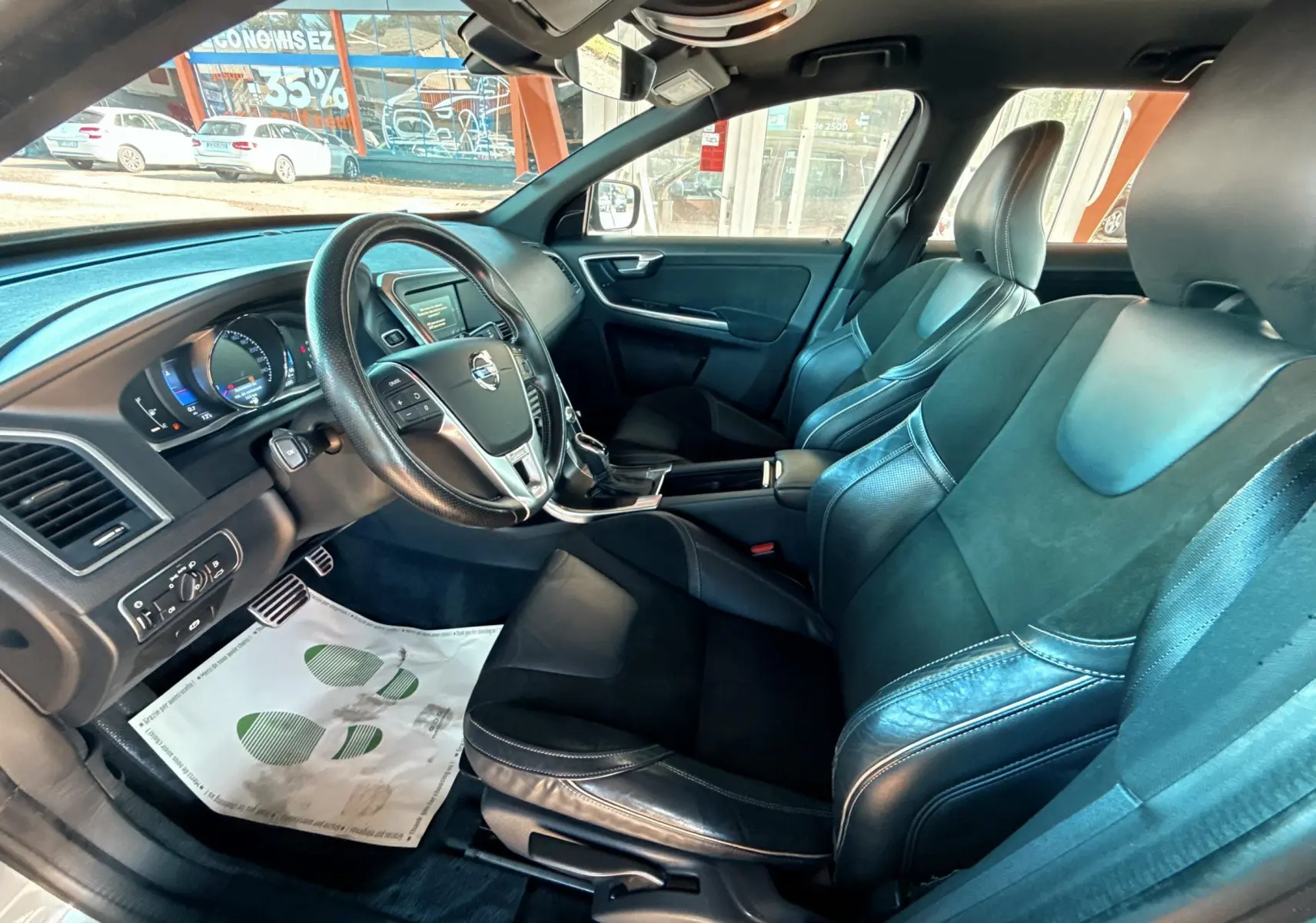 Vue intérieure côté conducteur du Volvo XC60 gris 2016, mettant en valeur le volant multifonction et les sièges cuir noir.