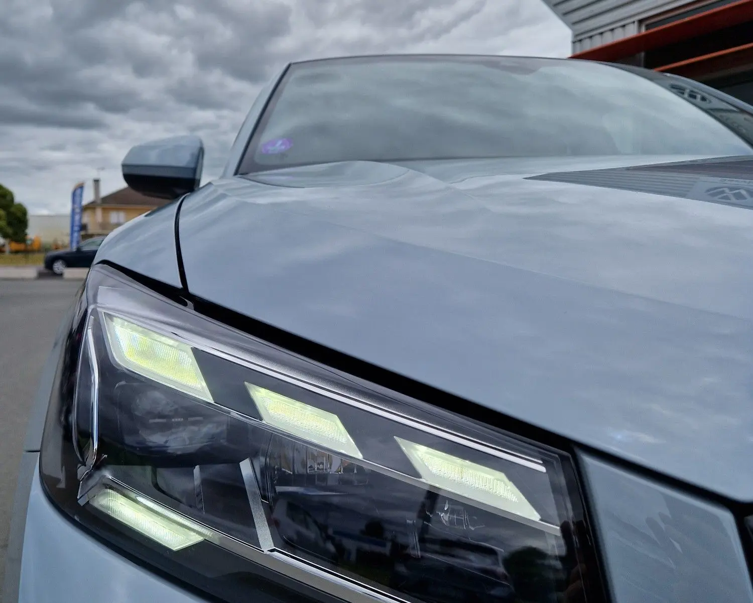 Gros plan sur le phare avant gauche de l'Audi Q2 35 TFSI 2023 gris flèche avec lignes LED distinctives sous ciel nuageux.