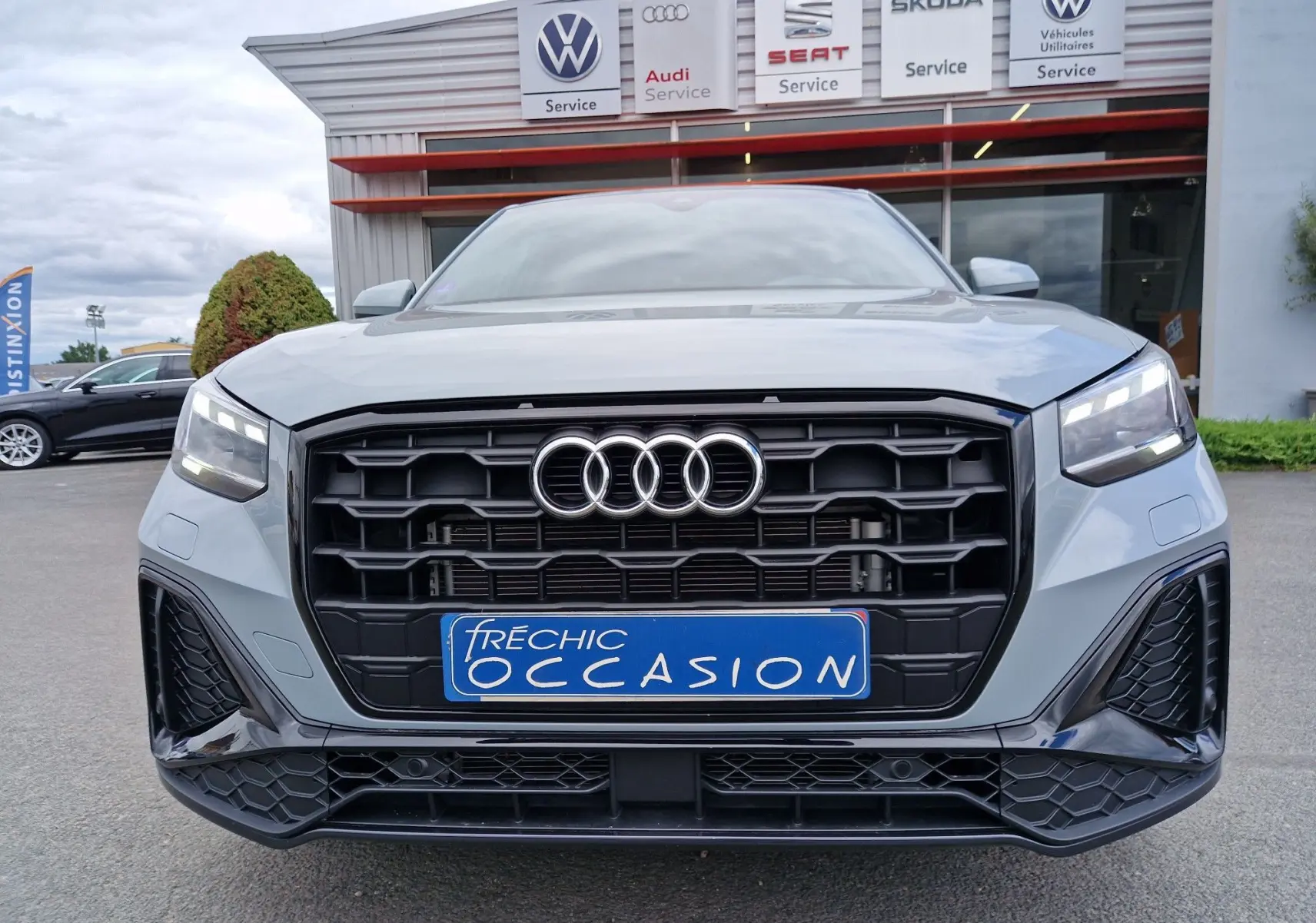 Vue frontale d’un Audi Q2 35 TFSI gris flèche avec calandre noire et phares LED allumés, finition S line visible.