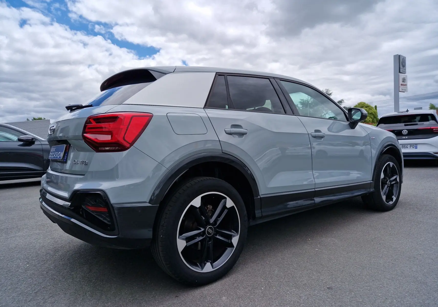 Audi Q2 35 TFSI 2023 gris flèche vue 3/4 arrière droit, toit blanc, jantes sport 18 pouces et vitres teintées.