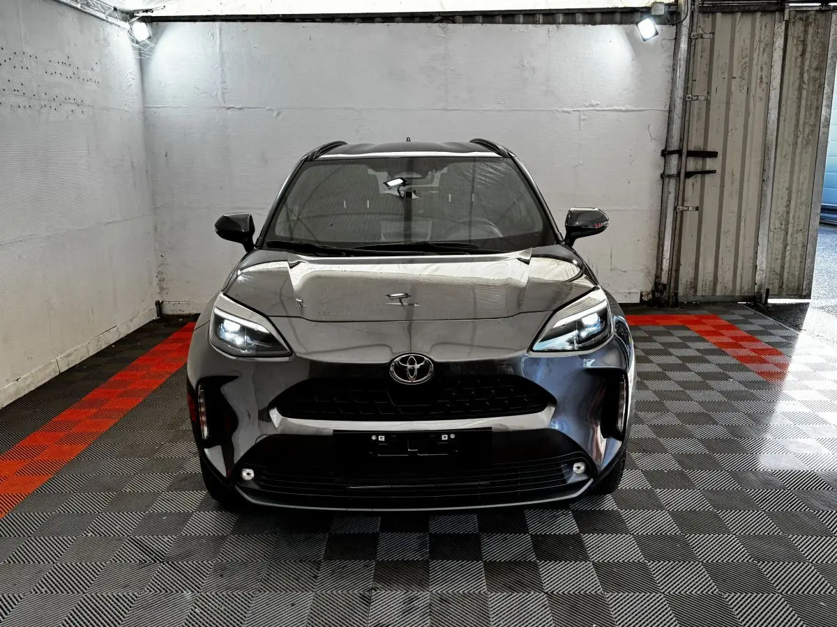 Vue frontale d'une Toyota Yaris Cross 1.5 Hybrid 130h Design Auto gris foncé avec phares LED allumés dans un garage.