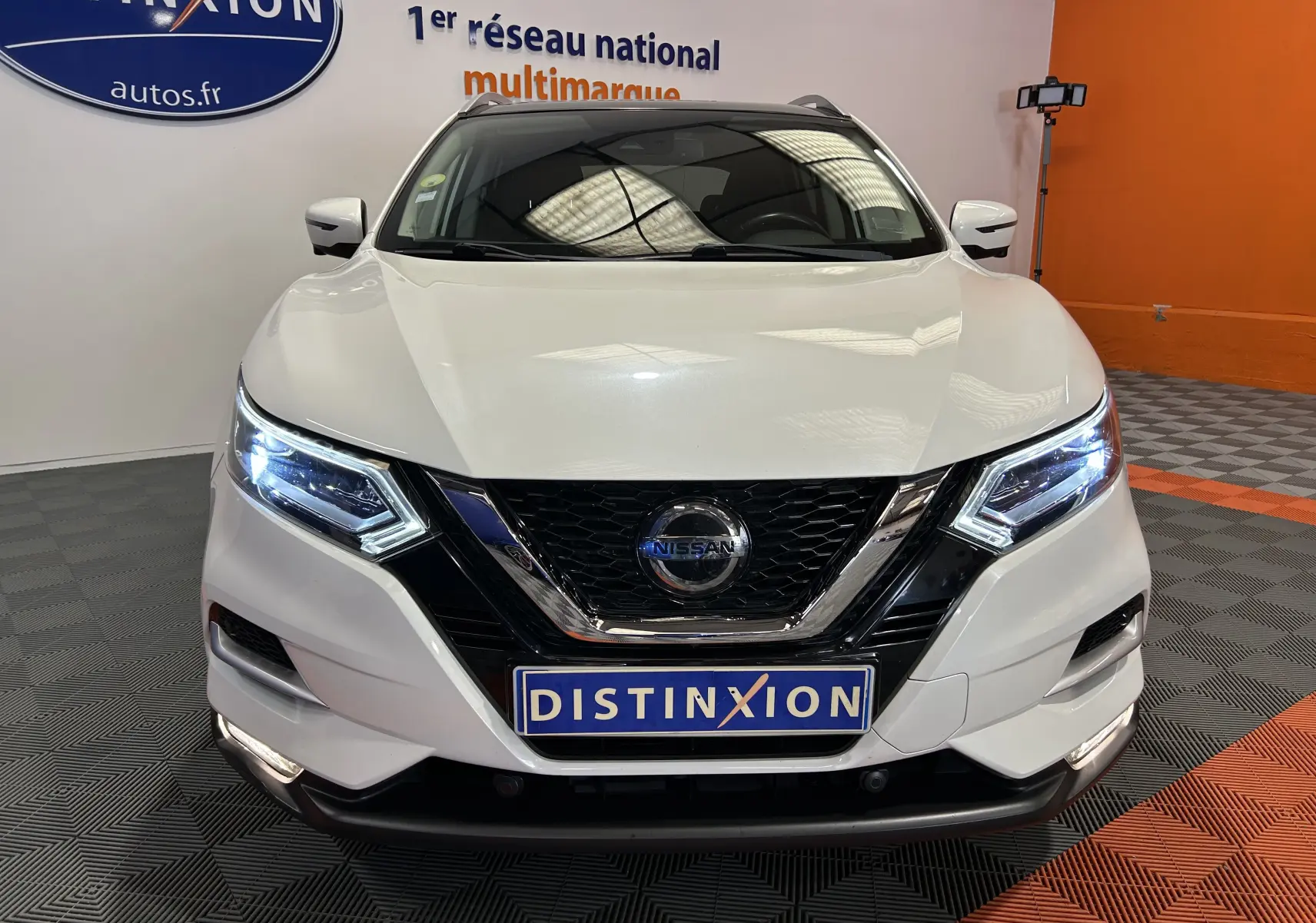 Vue de face d'un Nissan Qashqai blanc avec phares LED allumés et calandre noire brillante dans un showroom.