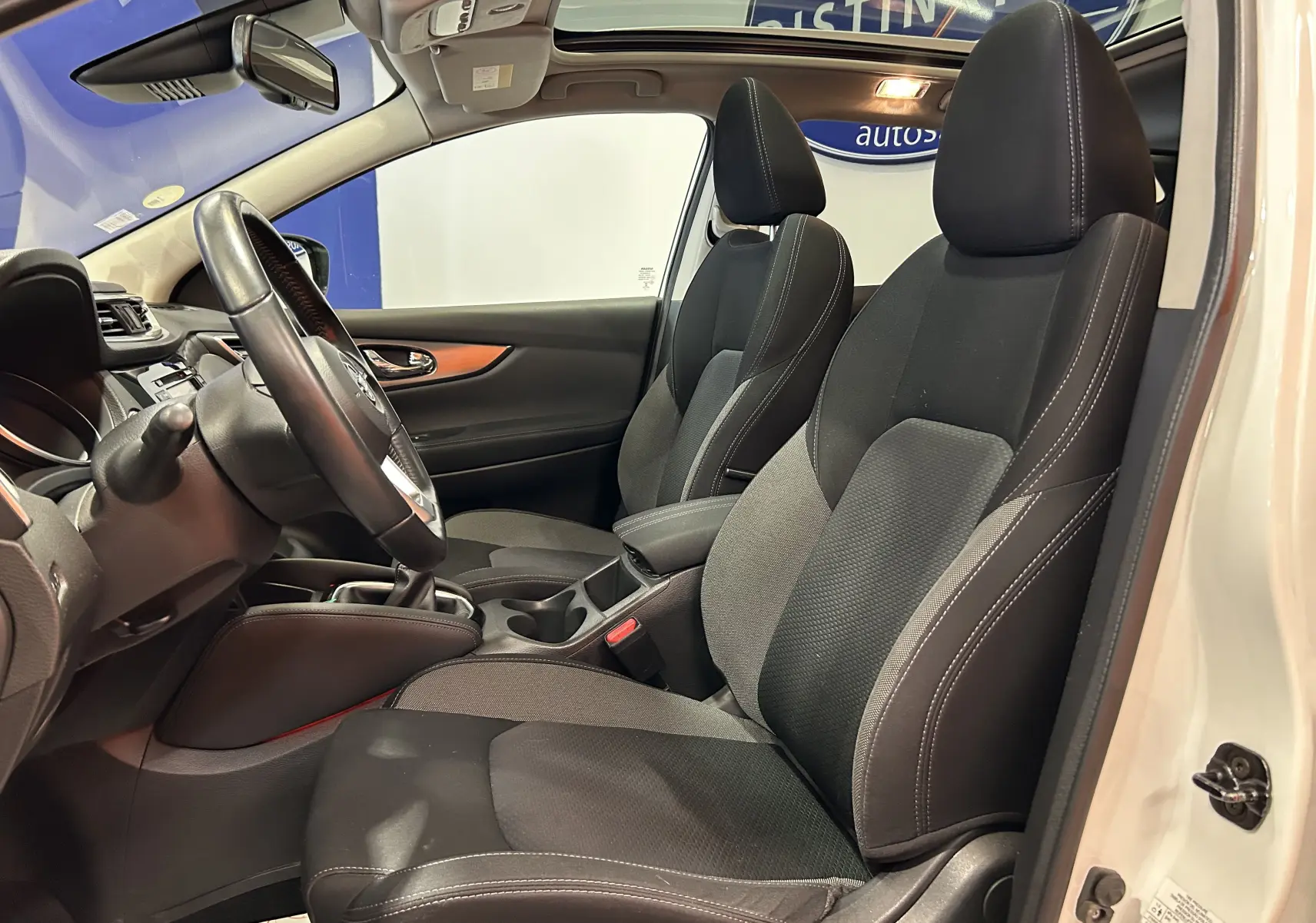 Vue intérieure côté conducteur du Nissan Qashqai blanc 2018, sièges tissu noir avec surpiqûres blanches et toit panoramique visible.