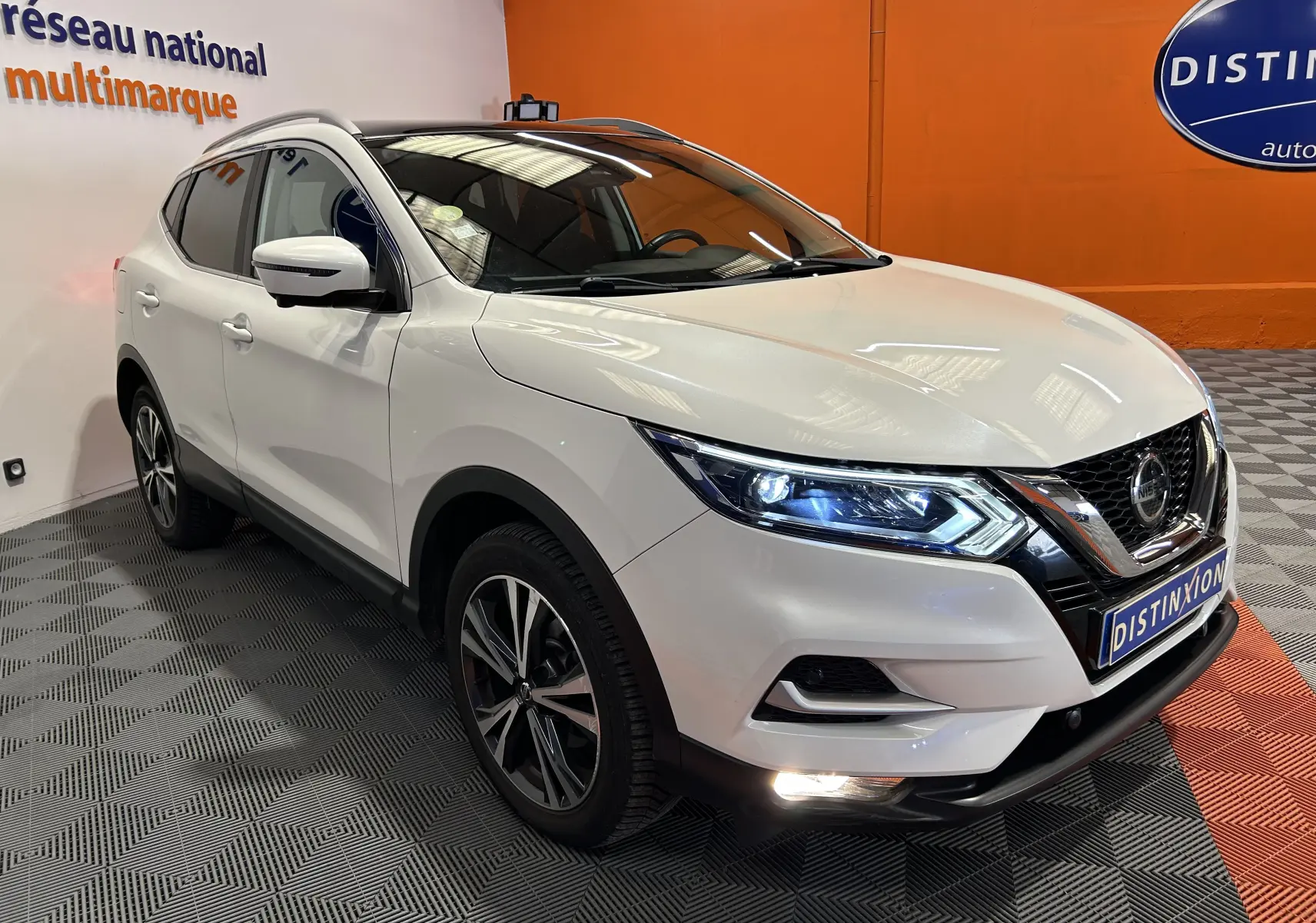Nissan Qashqai blanc en 3/4 avant droit, avec phares allumés et toit panoramique visible en intérieur showroom.