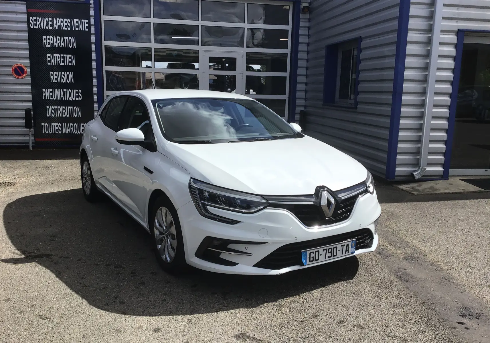 Renault Mégane IV Société blanche vue en 3/4 avant droit, stationnée devant un garage avec panneaux de service.