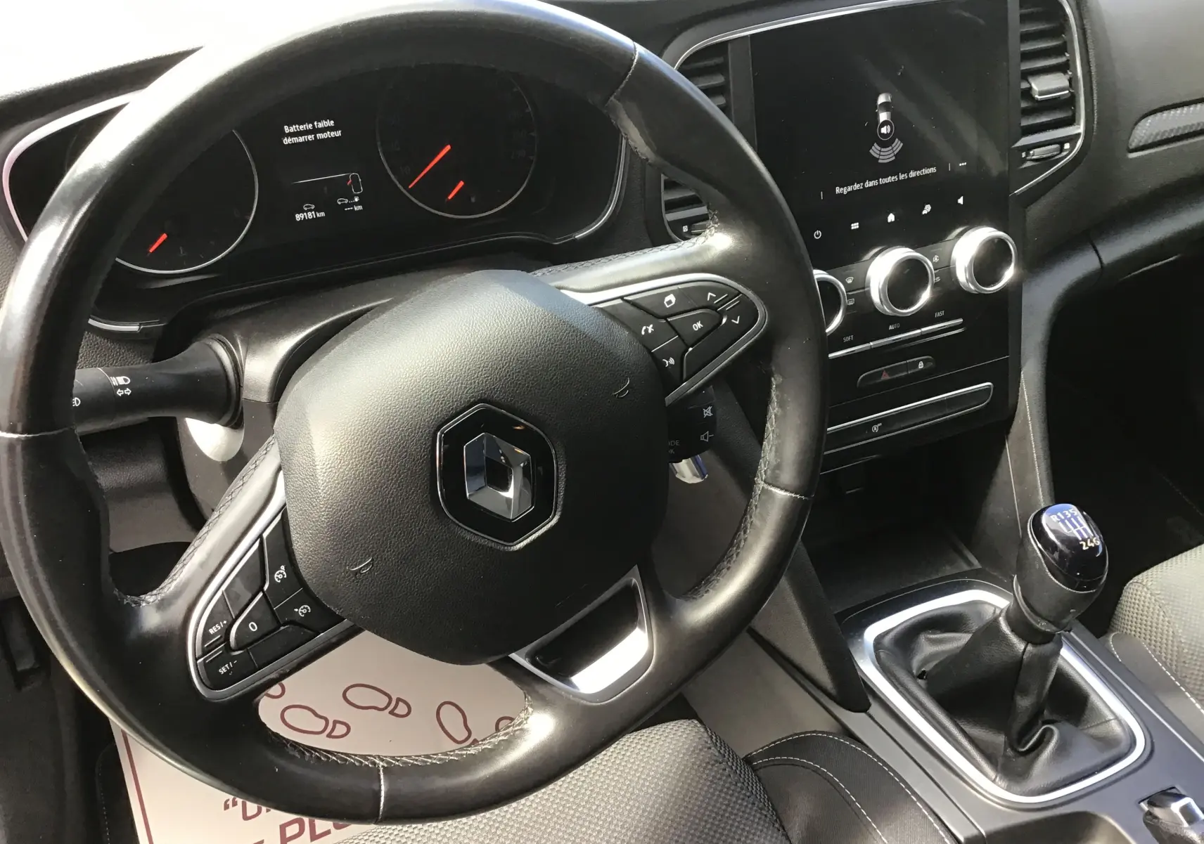 Vue intérieure centrée sur le volant noir et la console centrale avec levier de vitesse manuel d'une Renault Mégane IV Société blanche.