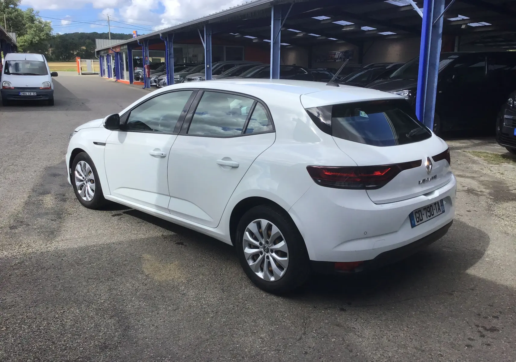 Vue 3/4 arrière droite d’une Renault Mégane IV Société blanche, avec feux arrière allongés et jantes alliage, stationnée en extérieur.