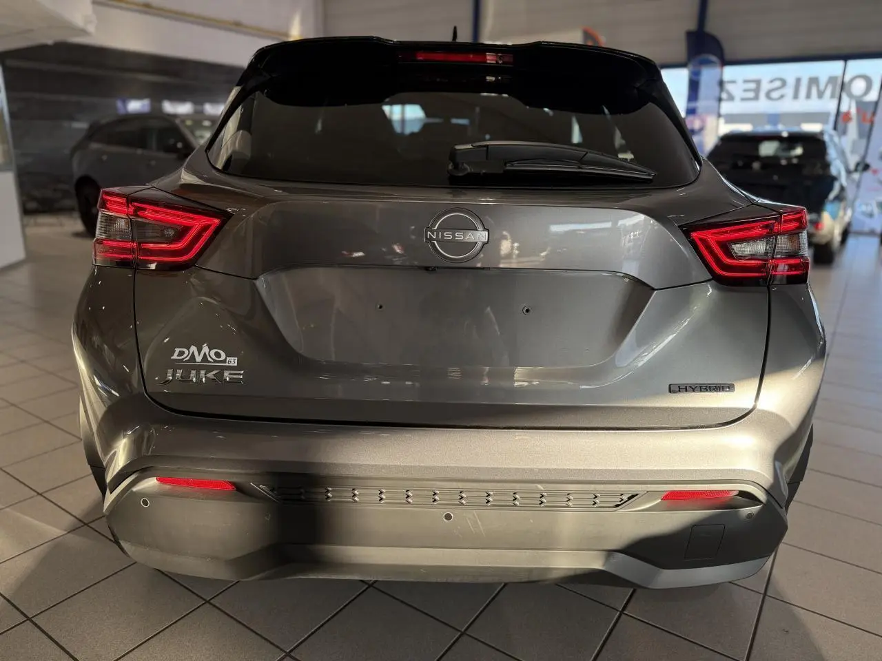 Vue arrière d'un Nissan Juke Hybrid gris métallisé avec feux arrière LED et logo Nissan visible en concession.