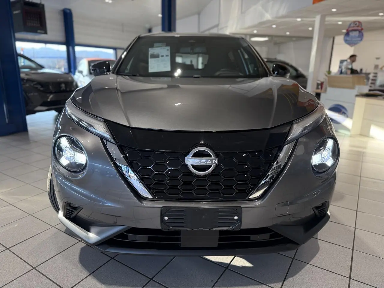 Vue frontale d'un Nissan Juke Hybrid gris métallisé en showroom avec calandre noire et phares LED ronds allumés.