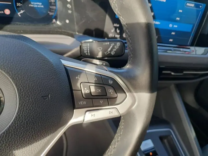 Gros plan sur les commandes gauche du volant cuir de la Volkswagen Golf noire, avec tableau de bord et écran tactile en arrière-plan.