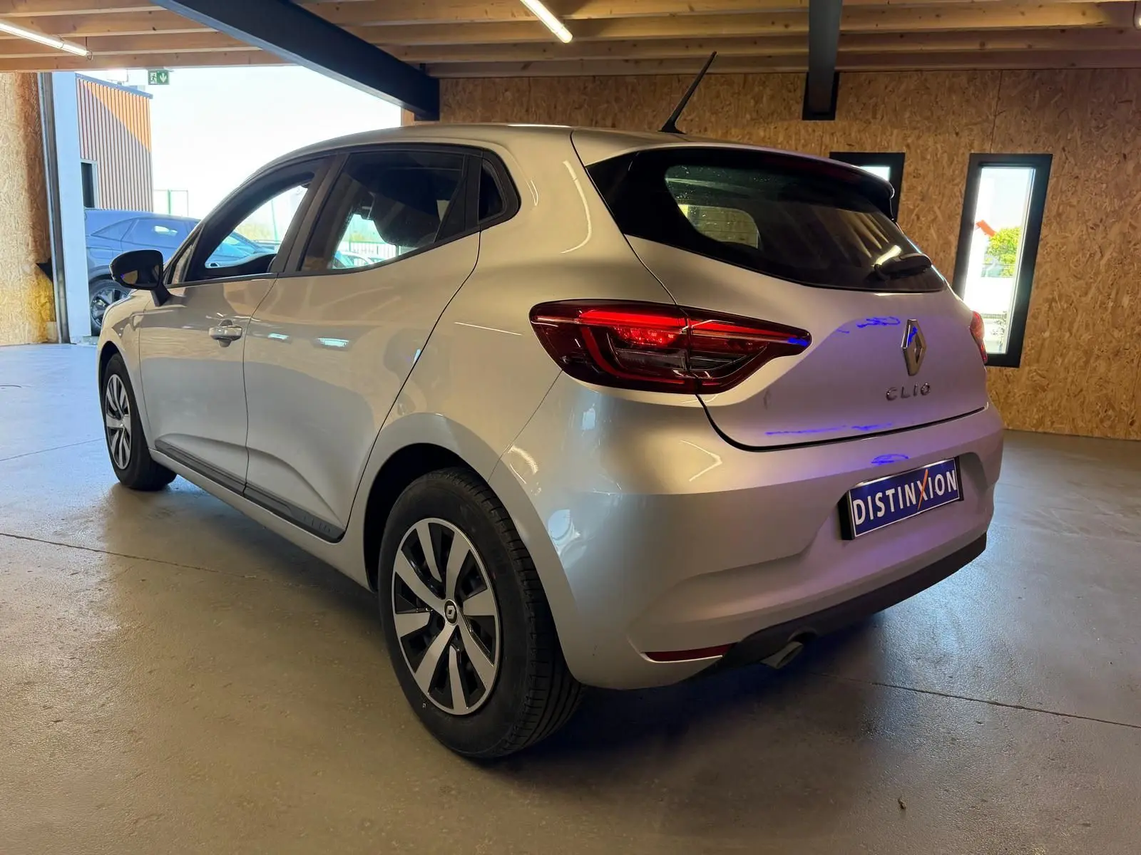 Vue 3/4 arrière droite d'une Renault Clio gris platine 2023 avec feux arrière LED et becquet dans un garage.