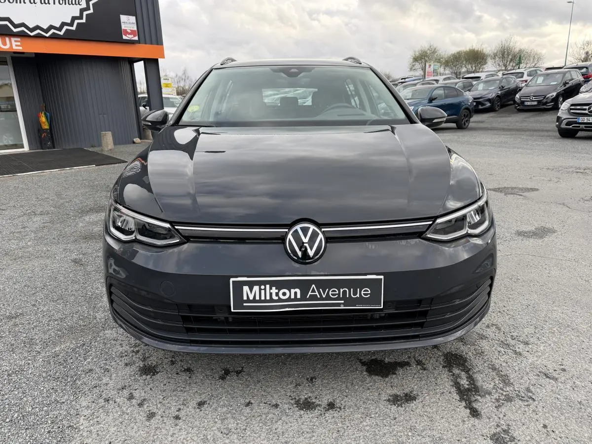 Vue frontale d'une Volkswagen Golf SW gris Urano avec calandre noire et logo VW centré sur un parking extérieur.