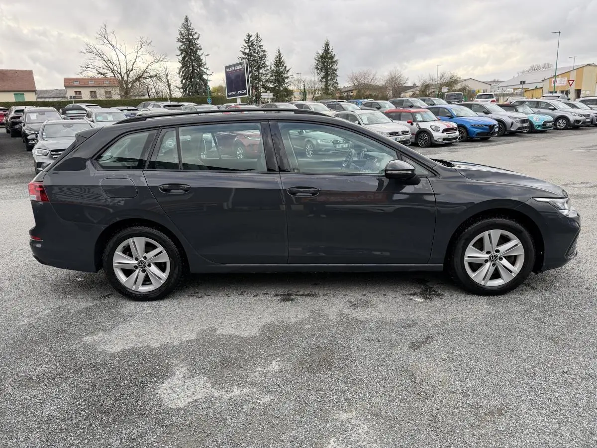 Vue latérale droite d'une Volkswagen Golf SW gris foncé stationnée sur un parking avec plusieurs autres véhicules en arrière-plan.