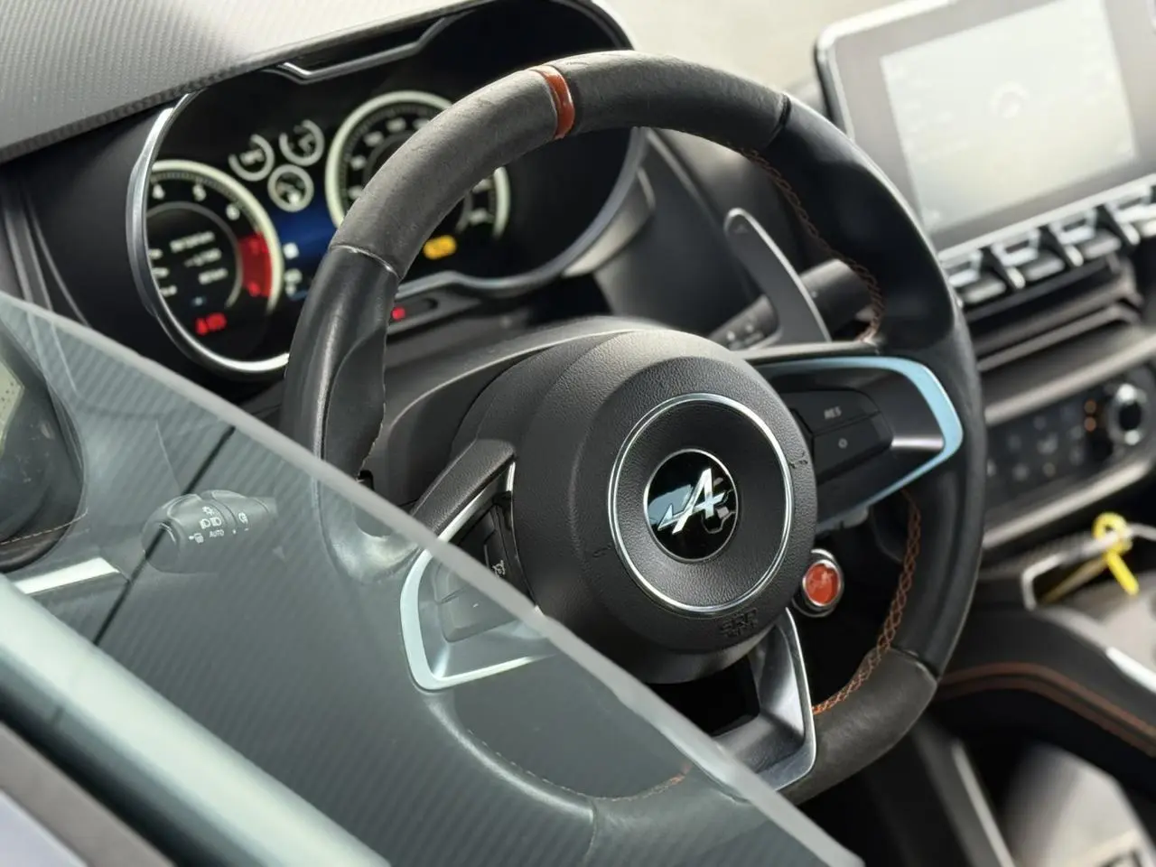 Gros plan sur le volant cuir noir avec surpiqûres orange de l'Alpine A110 1.8T 292 S, tableau de bord flou en arrière-plan.