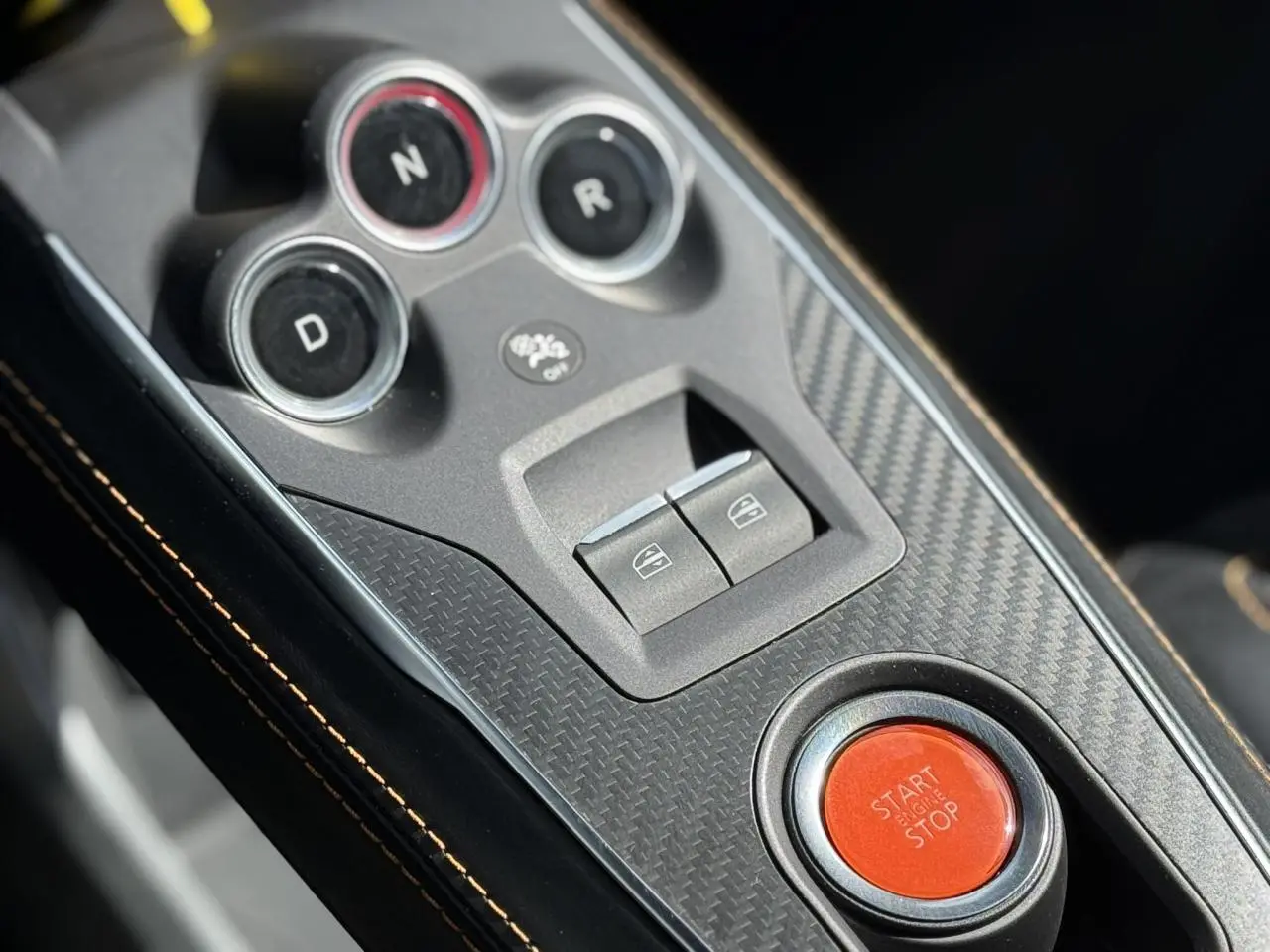 Gros plan sur la console centrale en carbone de l'Alpine A110 1.8T 292 S avec bouton start/stop rouge et surpiqûres orange.