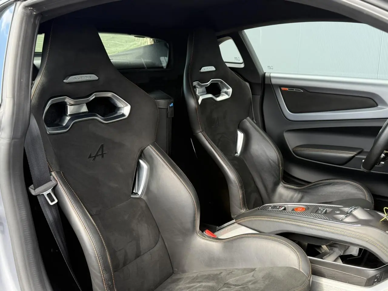 Vue intérieure côté gauche des sièges baquets noirs en cuir et Dinamica avec surpiqûres orange de l'Alpine A110 Gris Tonnerre Mat.