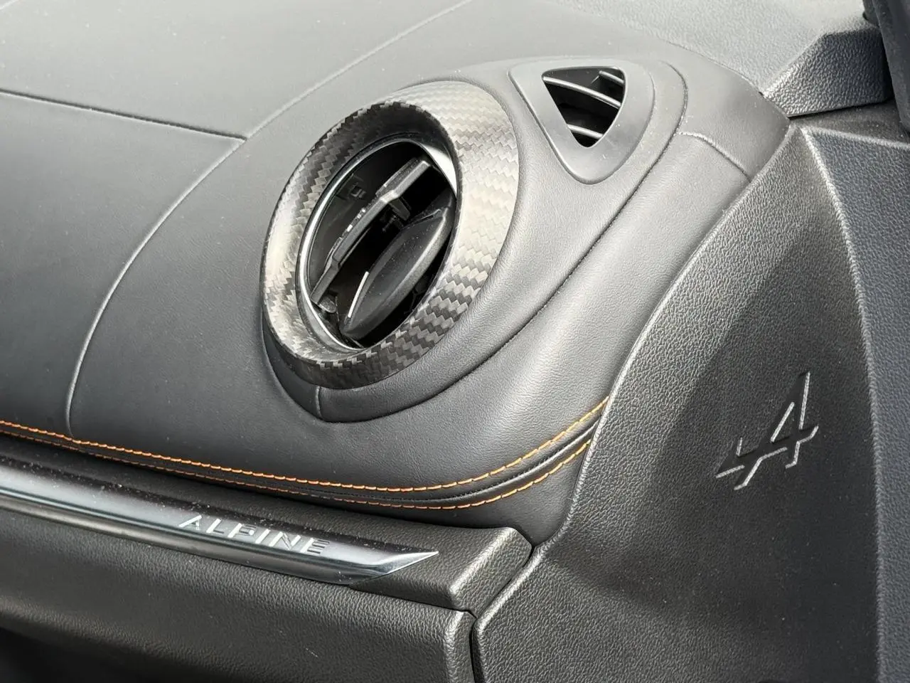 Détail intérieur côté passager de l'Alpine A110 gris mat, avec sortie d'air carbone et surpiqûres orange.